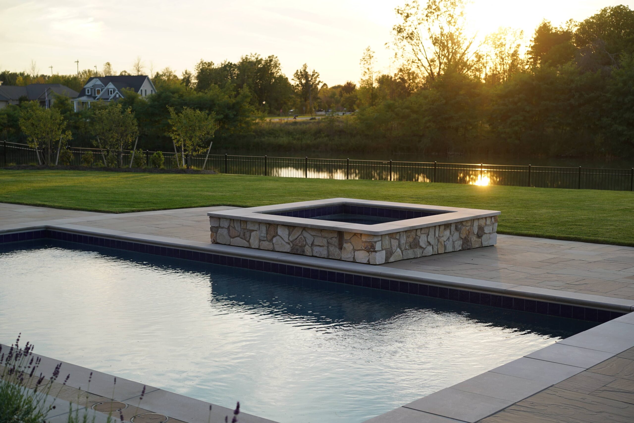 Backyard swimming pool with raised stone spa overlooking a landscaped lawn and waterfront at sunset.