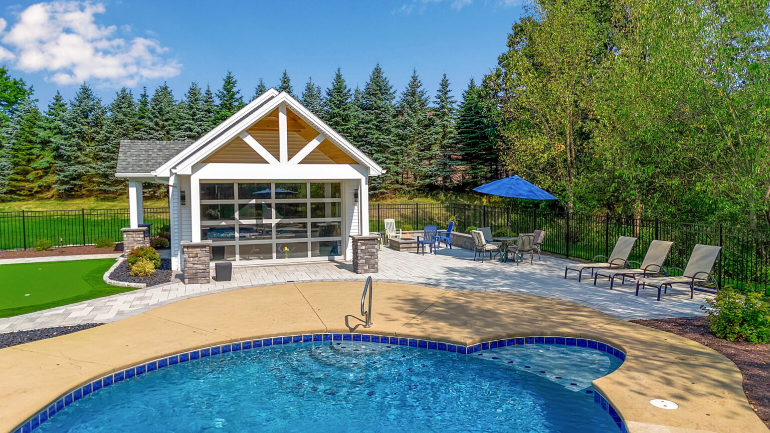 Backyard swimming pool with poolside pavilion, lounge chairs, and stone patio surrounded by landscaped greenery.