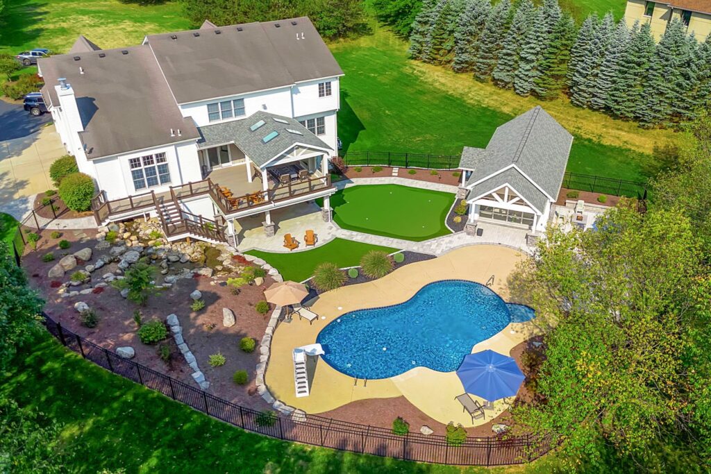 Aerial view of a landscaped backyard featuring a swimming pool, elevated deck, covered patio, putting green, and outdoor pavilion.