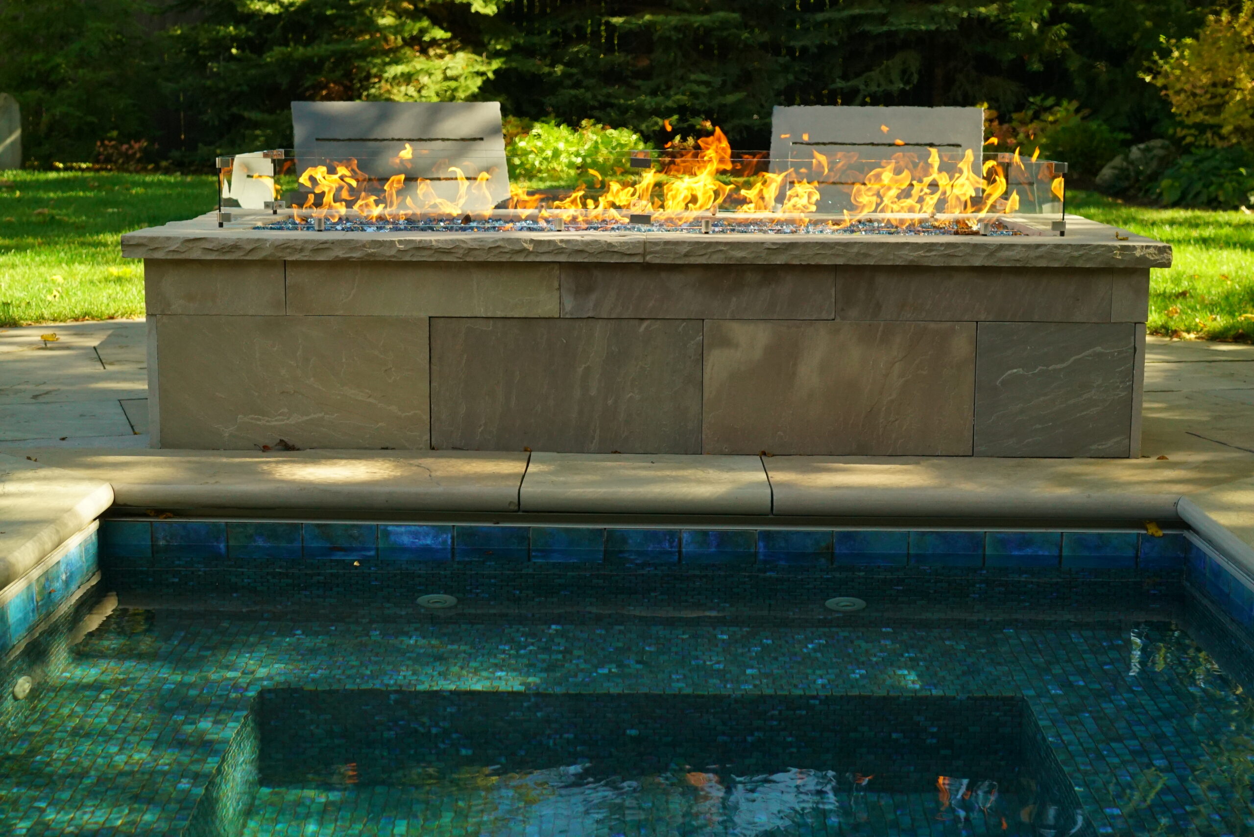 Modern linear fire feature beside a plunge pool with stone patio and lounge seating in a landscaped backyard.