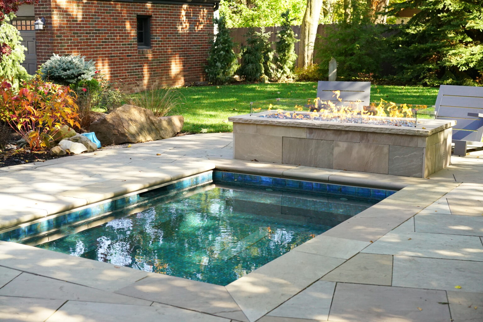 Modern backyard plunge pool with stone patio and built-in fire feature surrounded by landscaped greenery.