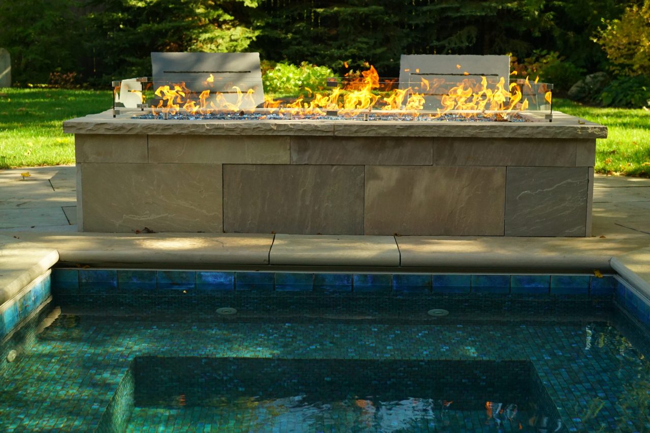 Modern linear fire feature beside a plunge pool with stone patio and lounge seating in a landscaped backyard.