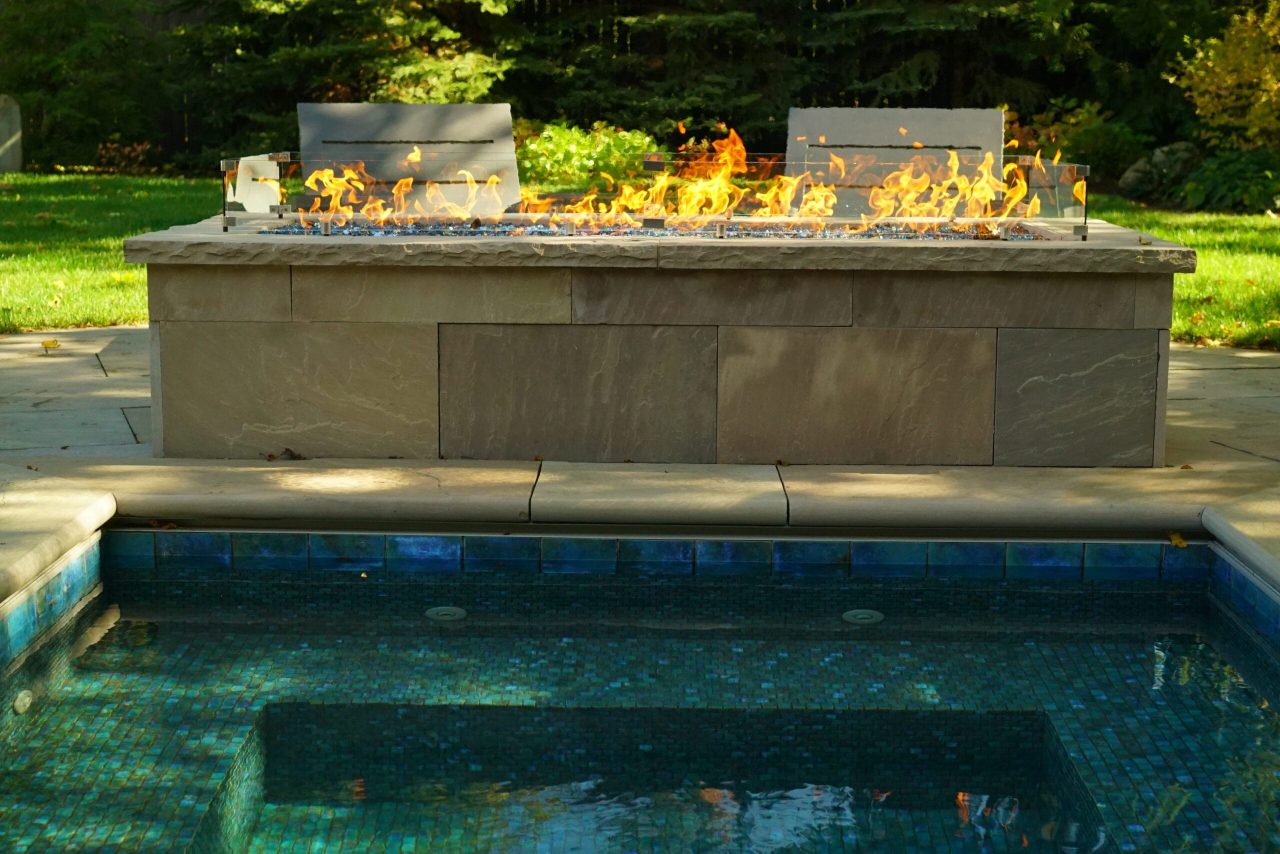 Modern linear fire feature beside a plunge pool with stone patio and lounge seating in a landscaped backyard.